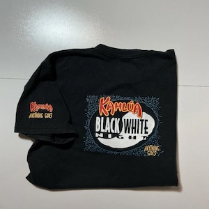 May include: Black t-shirt with the text "Kahlua Black White Night" in a white and orange design. The sleeve has the text "Kahlua Anything Goes" in orange. The shirt is folded on a white surface.