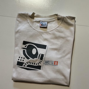 May include: Cream-colored t-shirt with a black graphic design and text. The design features a stylized image of a machine or device, with the text "AWS: ULTRX" and "SOVEREIGN SECT OF ALIEN WORKSHOP". The shirt is folded.