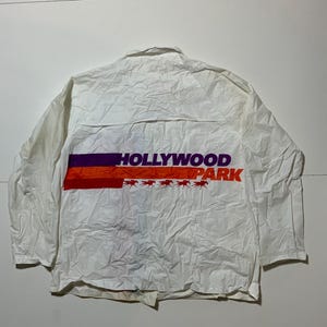 May include: White jacket with the text "HOLLYWOOD PARK" in purple and orange. The jacket has a horizontal stripe design across the chest. The text is above a row of horse silhouettes.