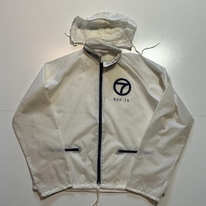 May include: A white windbreaker jacket featuring a hood, a dark blue front zipper, and two zippered pockets. The left chest displays a dark blue circular logo with the number seven inside, accompanied by the text KGO-TV printed in dark blue.
