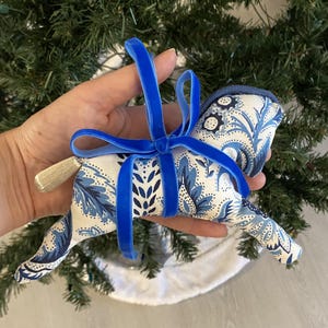 May include: A fabric horse Christmas ornament with a blue and white floral pattern. The ornament has a blue velvet ribbon bow and a gold tassel tail. The horse is held in front of a Christmas tree.
