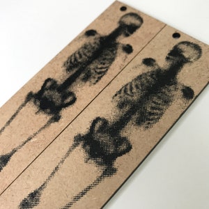 Könnte beinhalten: Zwei Holz-Lesezeichen mit schwarzen und weißen Skelett-Designs, die darauf gedruckt sind. Die Lesezeichen sind aus Naturholz gefertigt und haben ein rustikales Aussehen.