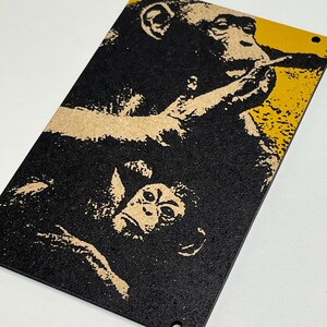 Könnte beinhalten: Ein rechteckiger schwarz-gelber Kunstdruck mit zwei Schimpansen. Der obere Schimpanse raucht, während der untere Schimpanse nach oben schaut. Der Druck hat ein strukturiertes Aussehen und enthält zwei kleine Löcher oben zum Aufhängen.