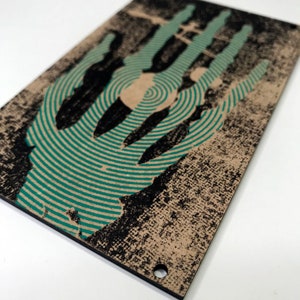 May include: A black and green wooden sign with a handprint design. The handprint is made up of concentric circles with a distressed look.