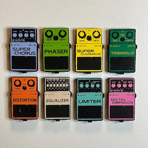 May include: Eight colorful guitar effect pedals are arranged in two rows. Each pedal has a different color and text label, including "Super Chorus," "Phaser," and "Super Overdrive." The pedals have knobs and input/output jacks.