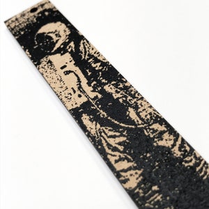 May include: A black and tan leather bookmark with a black and white graphic print of a person. The bookmark has a hole at the top for hanging.