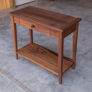 May include: A dark brown wooden console table with a single drawer and a lower shelf. The table has a simple, elegant design and is made of solid wood.