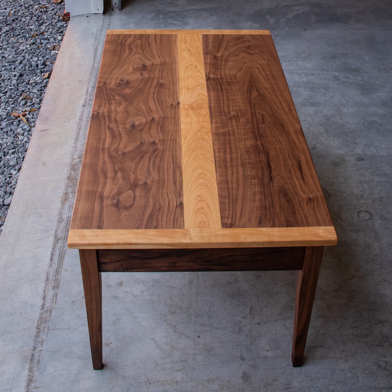 May include: A rectangular coffee table with a walnut wood top and a cherry wood inlay. The table has a simple design with four legs and a drawer.