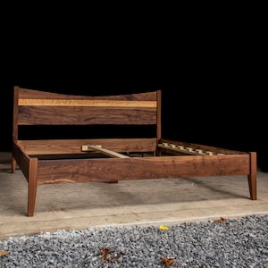 May include: A wooden bed frame with a curved headboard and a slatted base. The headboard is made of walnut wood with a natural finish. The frame is made of solid wood and has a dark brown finish.