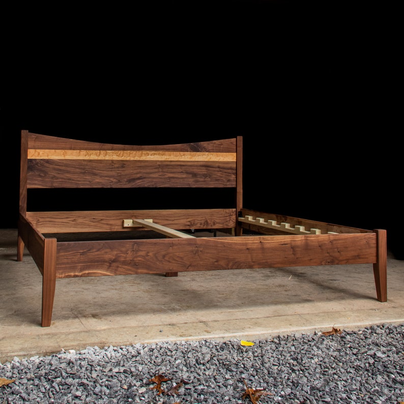 May include: A wooden bed frame with a curved headboard and a slatted base. The headboard is made of walnut wood with a natural finish. The frame is made of solid wood and has a dark brown finish.