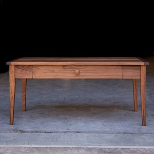 May include: A long, rectangular wooden table with a single drawer and four legs. The table is made of dark wood with a natural finish.