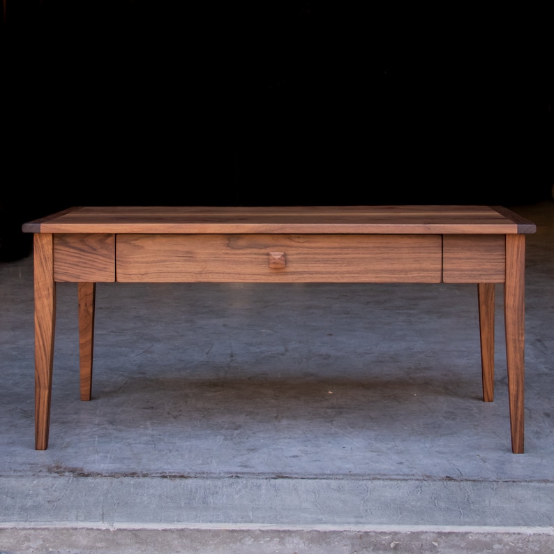 May include: A long, rectangular wooden table with a single drawer and four legs. The table is made of dark wood with a natural finish.