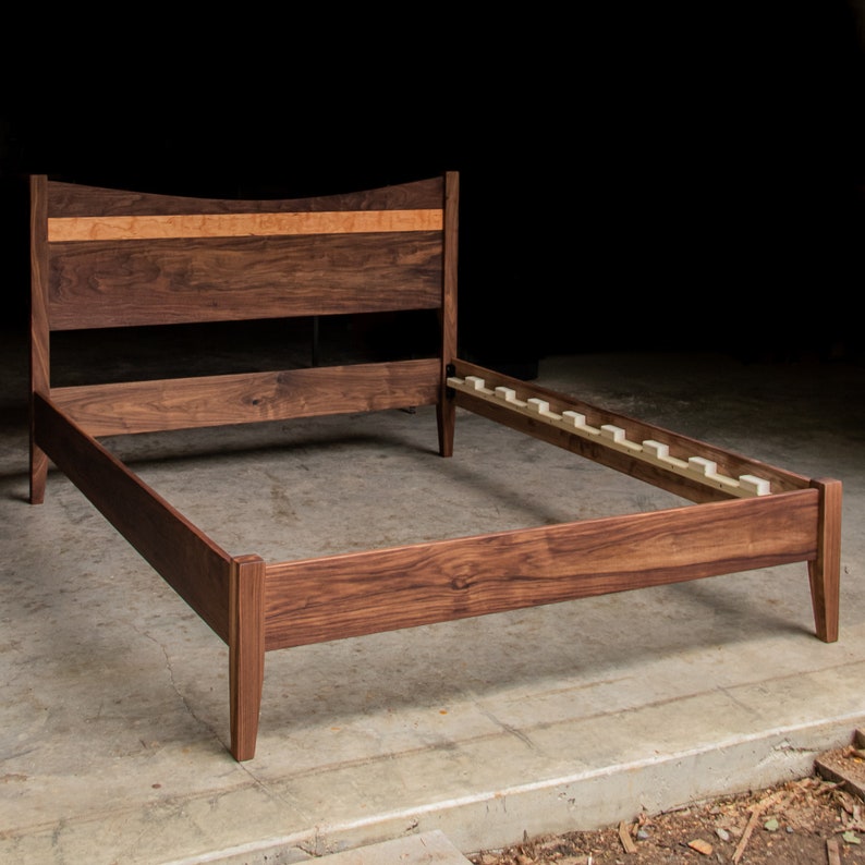 May include: A wooden bed frame with a curved headboard and a slatted base. The headboard is made of walnut wood with a maple inlay. The frame is a dark brown color.