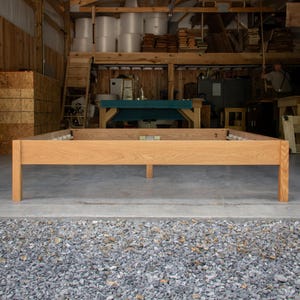 May include: A light brown wooden bed frame with a rectangular design. The frame has four sturdy legs and a central support beam. The bed frame is made of solid wood and appears to be unfinished. The frame is in a workshop setting.
