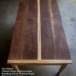 May include: A wooden table with a walnut top and ambrosia maple contrast stripe, breadboard ends, rails, and tapered legs.