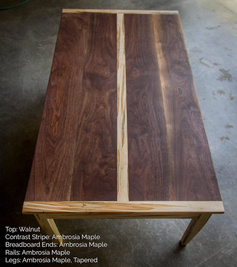 May include: A wooden table with a walnut top and ambrosia maple contrast stripe, breadboard ends, rails, and tapered legs.