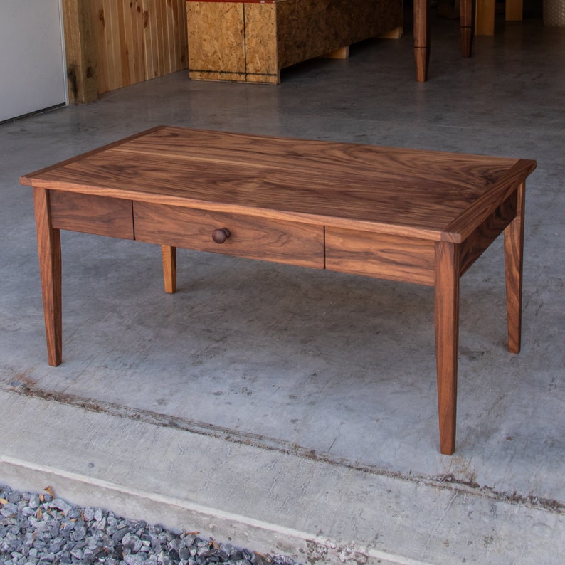 May include: A solid wood coffee table with a single drawer and a dark wood finish. The table has a simple, rectangular design with rounded edges and four legs.