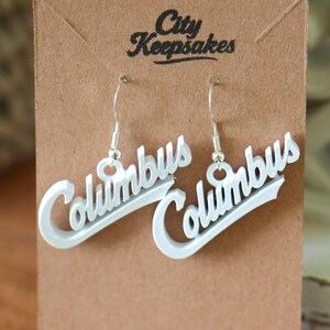 May include: White 'Columbus' script earrings on a brown card with 'City Keepsakes' text. The earrings are suspended from silver hooks. The background features a woven basket and a decorative white frame.