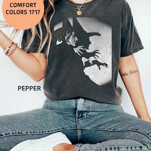 Puede incluir: Una camiseta de manga corta gris carbón con un estampado gráfico en blanco y negro de una bruja de perfil con sombrero puntiagudo y manos extendidas. La camiseta incluye el texto COMFORT COLORS 1717 y PEPPER impreso en la parte frontal.