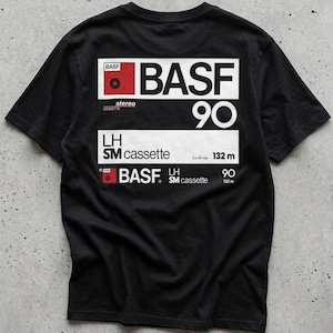 May include: A black short-sleeve t-shirt featuring a white graphic print on the back. The design mimics a BASF 90 cassette tape label with red and white accents, including the text BASF, 90, LH SM cassette, and 132 m in a rectangular layout.