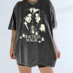 May include: Dark gray t-shirt featuring a graphic from the movie "Practical Magic." The design includes two women, candles, and the movie title in white lettering. The shirt has a vintage, worn look.