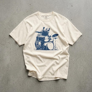 May include: A cream-colored short-sleeve t-shirt featuring a dark blue graphic print of a bear playing a drum set. The bear is positioned in the center of the chest, depicted with drumsticks, multiple drums, and cymbals in a detailed illustration.