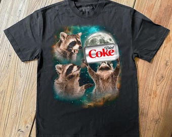 May include: Dark gray t-shirt featuring a graphic of three raccoons in space. One raccoon holds a Diet Coke can under a full moon. The shirt has the text "Killer Retro" at the top.