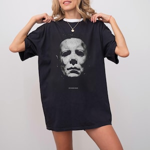 Könnte beinhalten: Schwarzes T-Shirt mit einem Graustufenbild einer Maske und dem Text "HE CAME HOME" darunter. Das Shirt ist dunkel und hat kurze Ärmel.