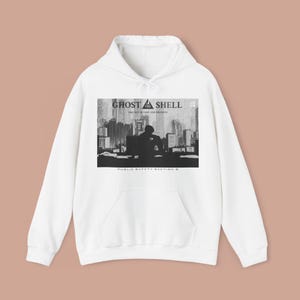 Puede incluir: Sudadera con capucha blanca con un diseño gráfico de "Ghost in the Shell". El diseño incluye el texto "GHOST IN THE SHELL" sobre una silueta de una persona frente a un paisaje urbano. Debajo está el texto "PUBLIC SAFETY SECTION 9".