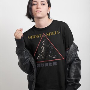 May include: Black t-shirt featuring the text "GHOST IN THE SHELL" in gold lettering above a red triangle graphic. The graphic includes a silhouette of a figure. Below the triangle is Japanese text.