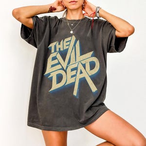 May include: Dark grey t-shirt featuring the text "THE EVIL DEAD" in a distressed, vintage style. The graphic has a blue shadow effect. The shirt has short sleeves and a relaxed fit.