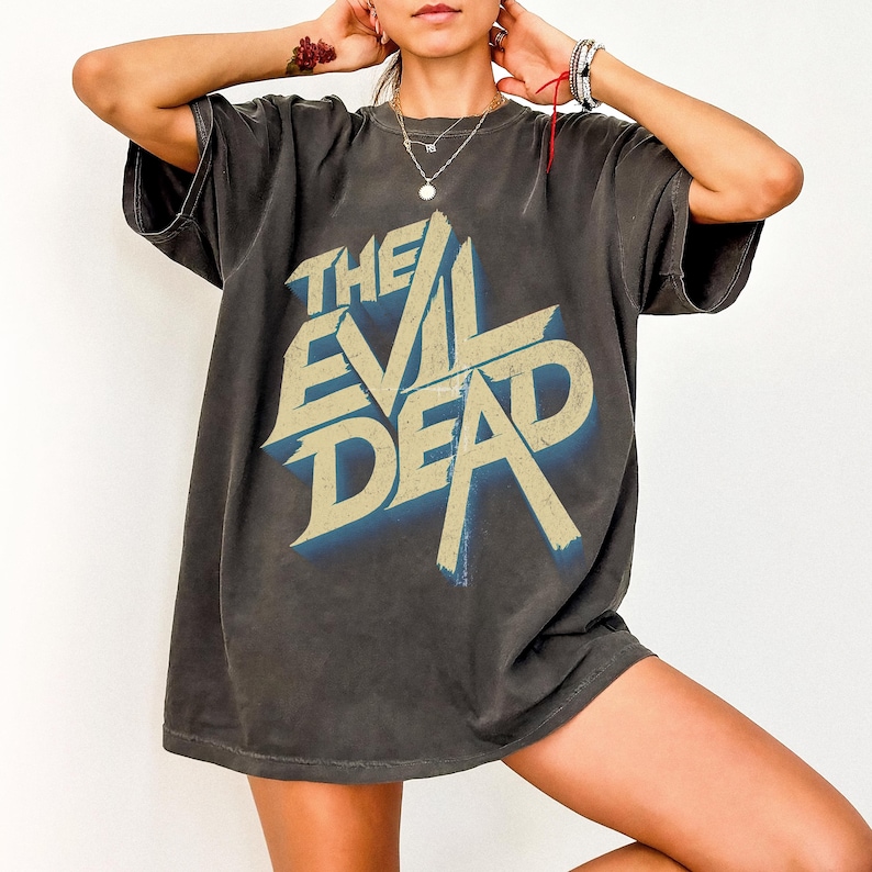 May include: Dark grey t-shirt featuring the text "THE EVIL DEAD" in a distressed, vintage style. The graphic has a blue shadow effect. The shirt has short sleeves and a relaxed fit.