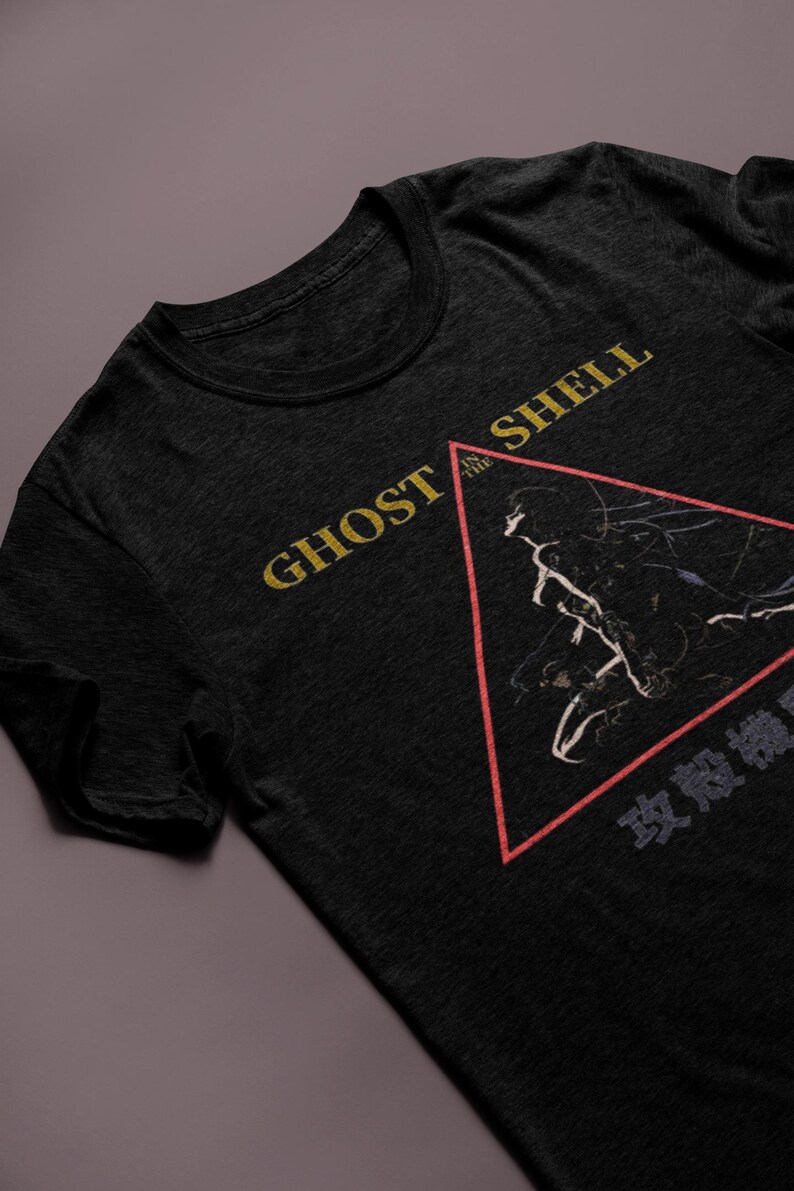 May include: Dark gray t-shirt featuring the text "GHOST IN THE SHELL" in gold lettering. A red-outlined triangle contains a black and white graphic. Japanese text is below the triangle. The shirt is made of a soft material.
