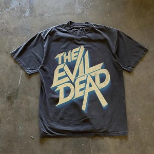 May include: A dark gray t-shirt featuring the text "THE EVIL DEAD" in a distressed, stylized font with a blue shadow effect. The shirt is made of a soft material and has a classic crew neck.