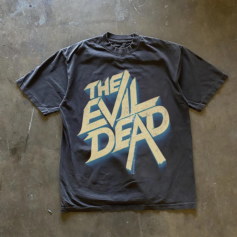 May include: A dark gray t-shirt featuring the text "THE EVIL DEAD" in a distressed, stylized font with a blue shadow effect. The shirt is made of a soft material and has a classic crew neck.