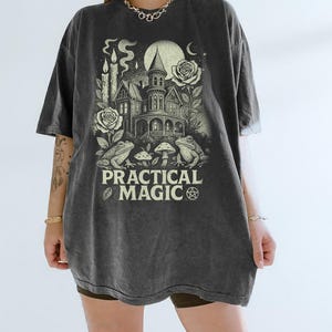 Puede incluir: Camiseta gris oscuro con un gráfico que presenta una casa embrujada, rosas, ranas y el texto "PRACTICAL MAGIC". El diseño incluye una luna llena, velas y setas, creando un tema místico.