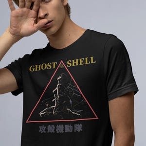 Może przedstawiać: Czarny t-shirt z napisem "GHOST IN THE SHELL" w kolorze złotym i czerwonym trójkątem z grafiką. Japoński tekst znajduje się pod trójkątem. Koszulka wykonana jest z miękkiego materiału.