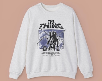 May include: A light gray sweatshirt featuring a graphic from the movie "The Thing." The design includes the film's title and a stylized image of a creature. The text "ANYTIME. ANYWHERE. ANYONE. MAN IS THE WARMEST PLACE TO HIDE." is also printed on the sweatshirt.
