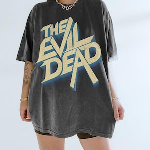 May include: Dark gray t-shirt with the text "THE EVIL DEAD" in a distressed, vintage style. The text is in a light yellow color with a blue shadow effect. The shirt has a relaxed fit.