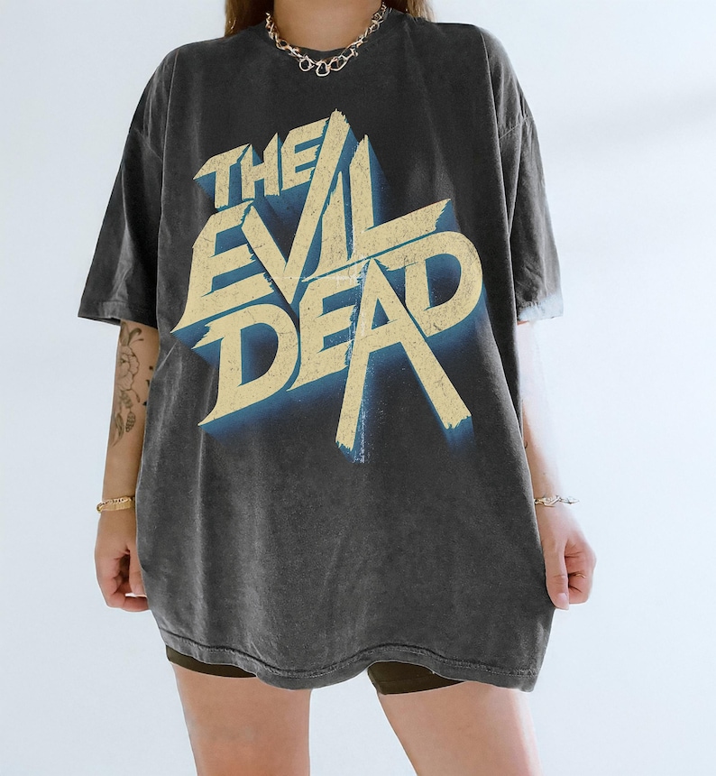 May include: Dark gray t-shirt with the text "THE EVIL DEAD" in a distressed, vintage style. The text is in a light yellow color with a blue shadow effect. The shirt has a relaxed fit.