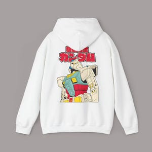Könnte beinhalten: Ein weißer Kapuzenpullover mit einem Grafikdruck auf der Rückseite. Das Design zeigt einen großen, mehrfarbigen Roboter in Creme, Blau, Rot und Gelb, mit japanischen Schriftzeichen in kräftigem Rot, die über dem Roboter platziert sind.