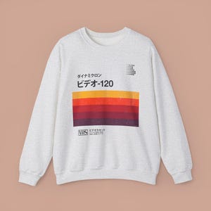 Puede incluir: Sudadera gris claro con un diseño retro de cinta VHS. El diseño incluye rayas horizontales en tonos naranja, rojo y marrón, con texto japonés arriba y abajo. La sudadera tiene cuello redondo y mangas largas.
