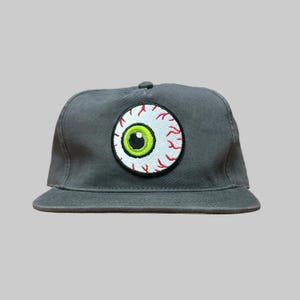 May include: A charcoal gray baseball cap featuring a circular embroidered patch on the front. The patch depicts a realistic human eyeball with a bright green iris, a black pupil, and red blood vessels radiating across the white sclera.