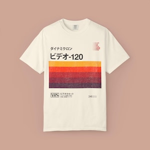Puede incluir: Camiseta de color crema con un diseño retro que recuerda a una etiqueta de cinta VHS. El diseño incluye texto japonés, un bloque de color con rayas naranjas, rojas y marrones, y el logotipo de VHS. La camiseta tiene cuello redondo clásico y mangas cortas.