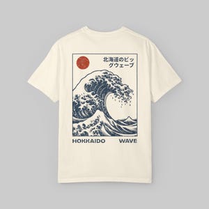 May include: Cream-colored t-shirt featuring a navy blue wave graphic with a red sun and Japanese text. The text reads "HOKKAIDO WAVE". The design is a vintage-style illustration.