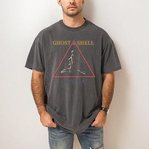 May include: Dark gray t-shirt with the text "GHOST IN THE SHELL" in gold letters above a red triangle with a white graphic. Below the triangle is Japanese text. The shirt is worn by a person.
