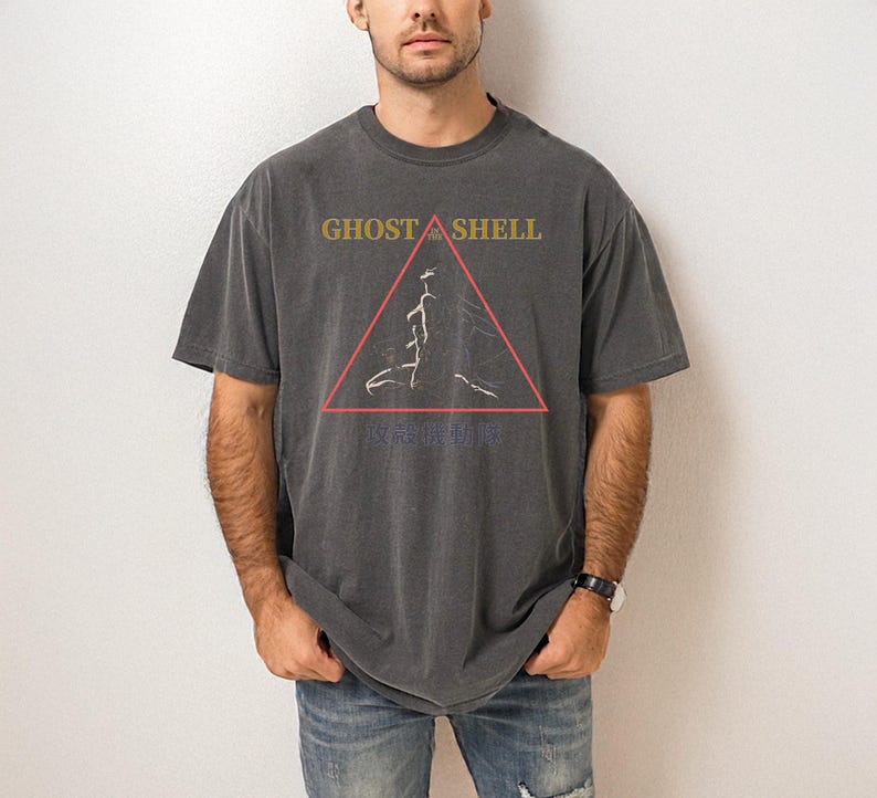 May include: Dark gray t-shirt with the text "GHOST IN THE SHELL" in gold letters above a red triangle with a white graphic. Below the triangle is Japanese text. The shirt is worn by a person.
