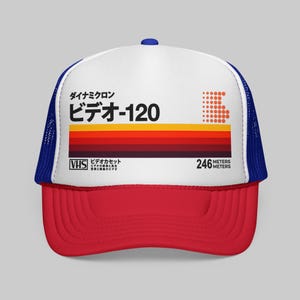 Peut inclure: Une casquette trucker blanche avec une visière rouge et un filet bleu à l'arrière. Le panneau avant présente un motif rappelant une étiquette de cassette VHS avec du texte japonais, le nombre 120, des rayures rouges et orange et 246 METERS.