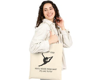 May include: A cream-colored tote bag featuring a black silhouette of a gymnast in a split leap. The text on the bag reads "If you don't leap, you'll never know what it's like to fly."