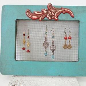 May include: A turquoise blue wooden frame with a wire mesh insert displays three pairs of dangle earrings. The earrings feature gold filigree, red and blue beads.
